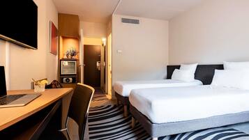 Habitación de hotel con escritorio, dos camas, televisor y puerta que da a un pasillo.