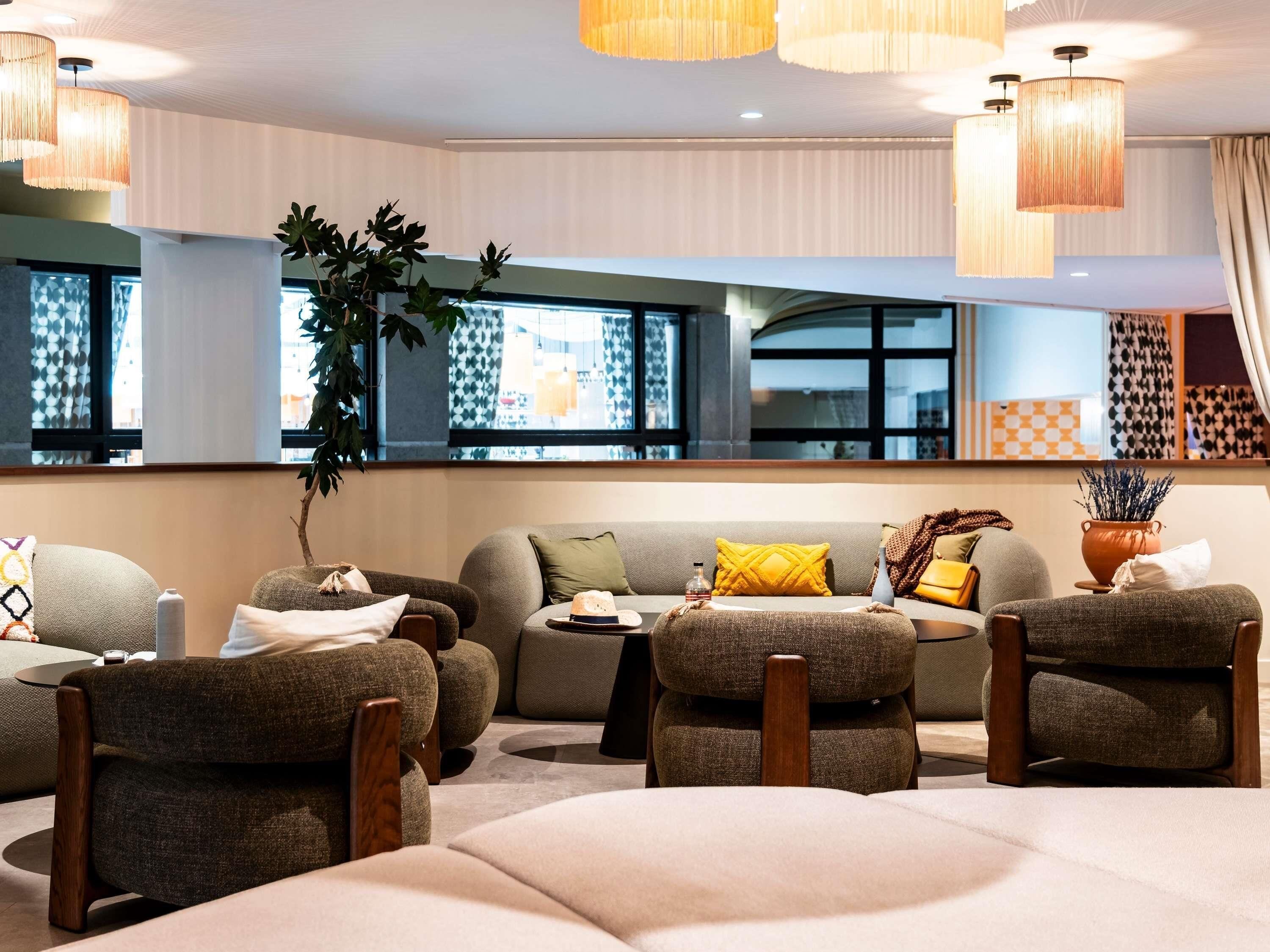 A modern lounge area with a sofa, armchairs, and a coffee table. There are large windows with patterned curtains, a potted plant, and a decorative vase.