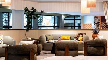 A modern lounge area with a sofa, armchairs, and a coffee table. There are large windows with patterned curtains, a potted plant, and a decorative vase.