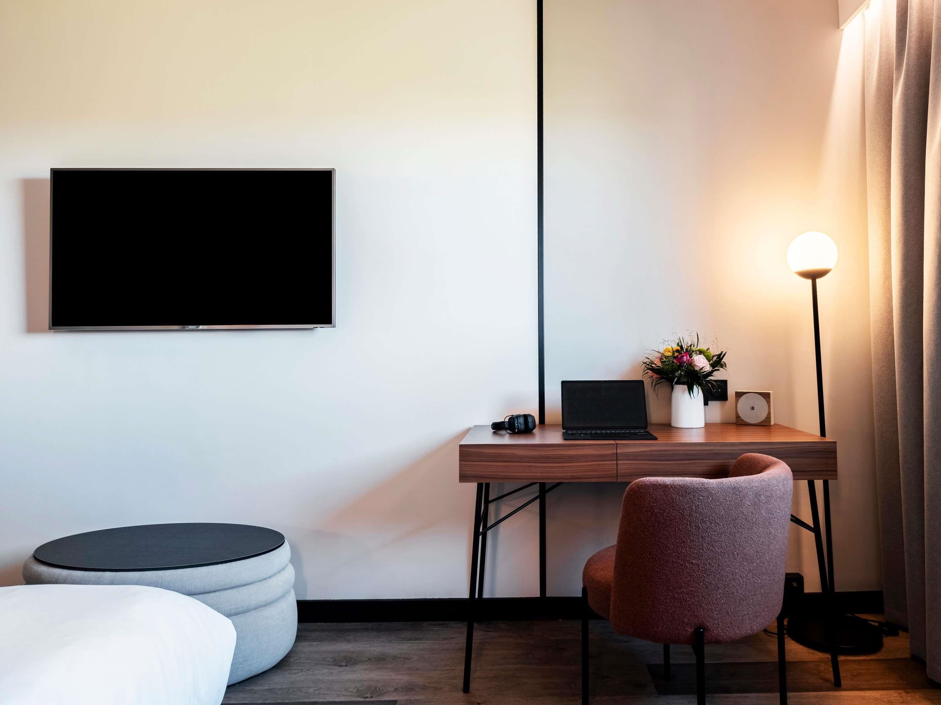A modern hotel room with a flat-screen TV, a desk with a laptop, a lamp, and a chair.