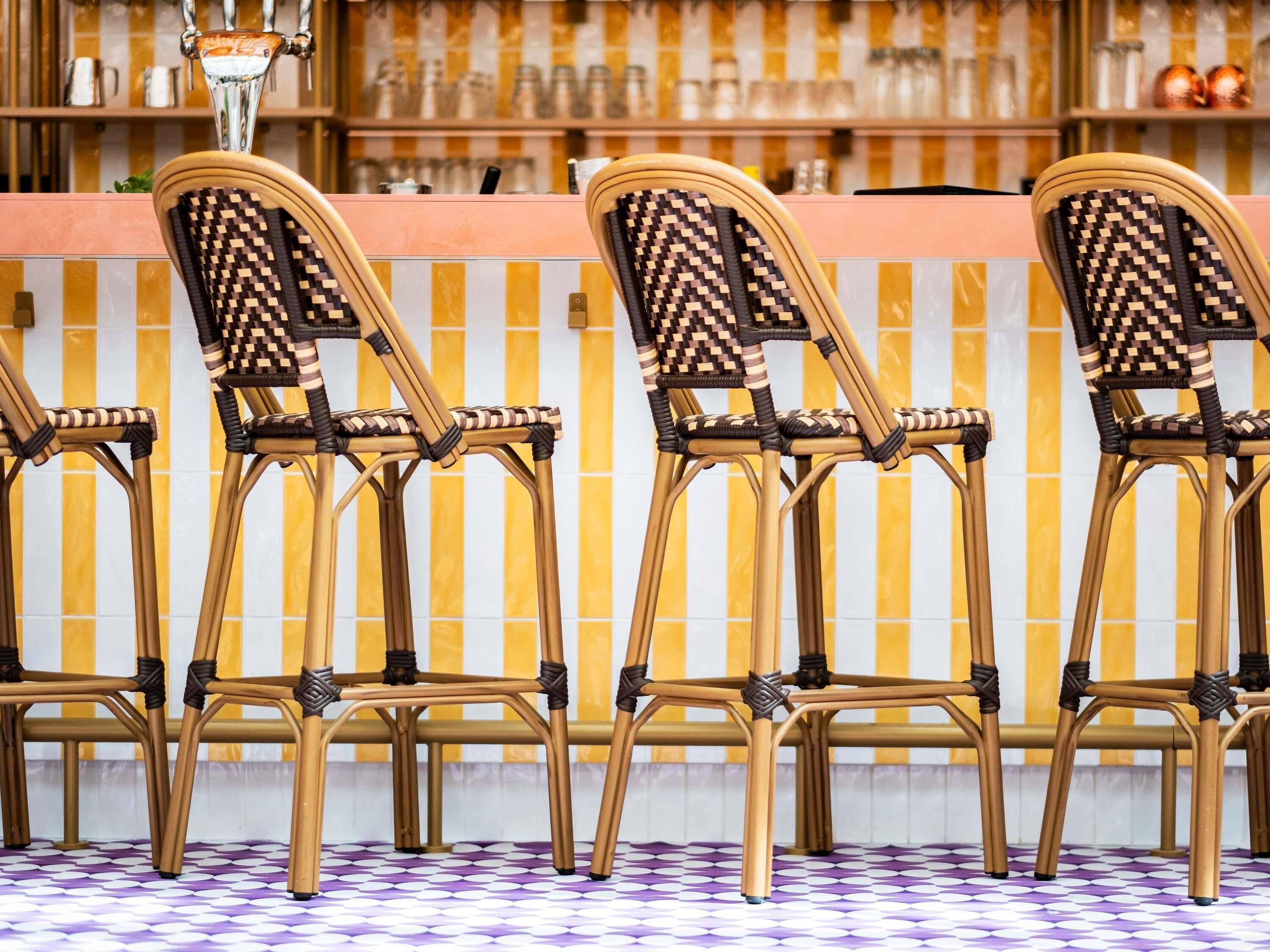 A row of wicker chairs with woven backs and metal frames, arranged in front of a bar area with a tiled backsplash.