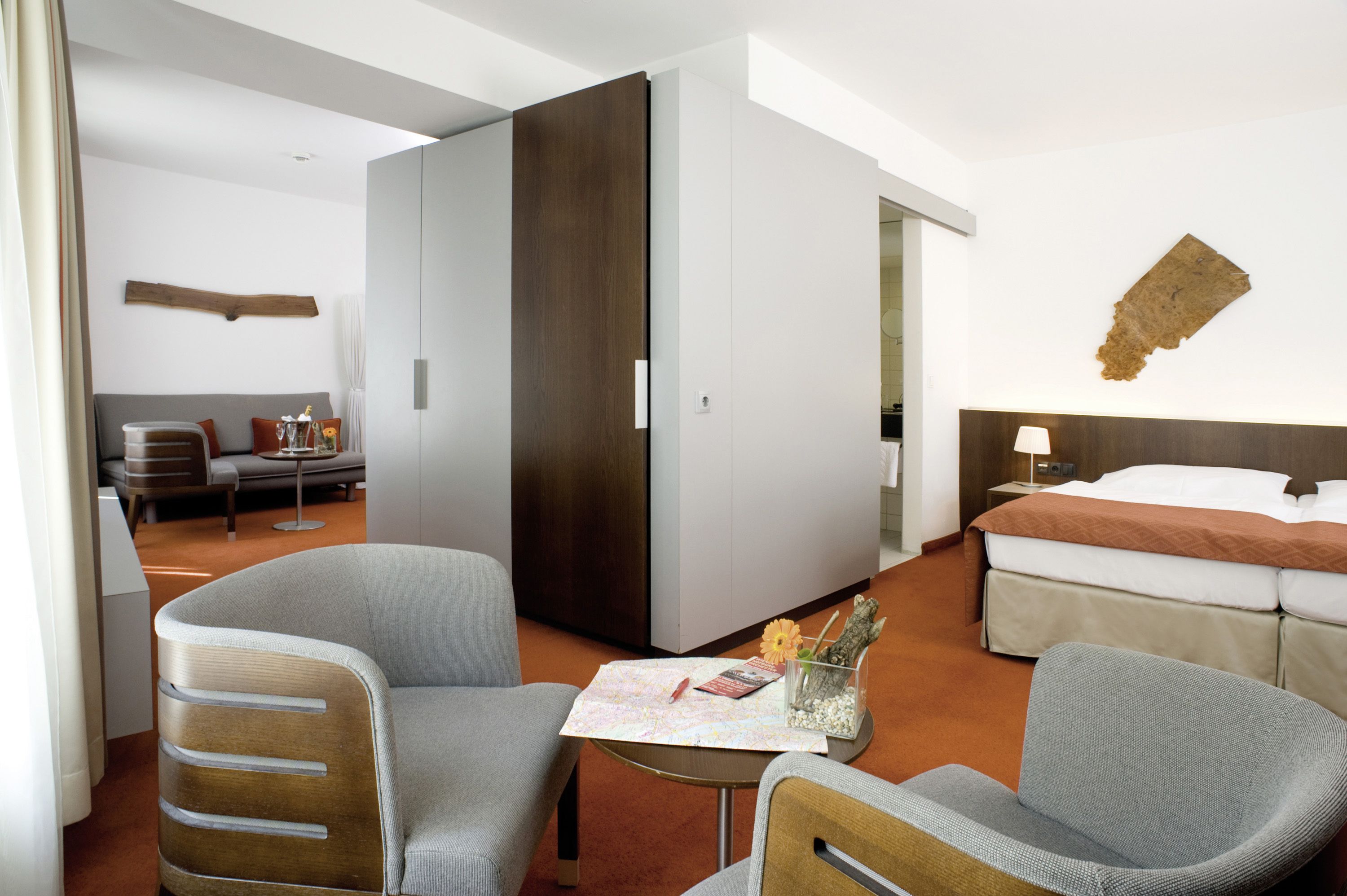 A hotel room with a bed, two armchairs, a small table with a vase and book, a wooden cabinet, and a wall-mounted artwork.