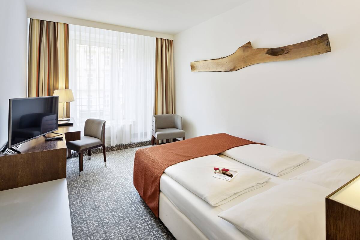 A hotel room with a bed, a desk with a TV, a chair, a window with curtains, and a wall-mounted wooden piece.