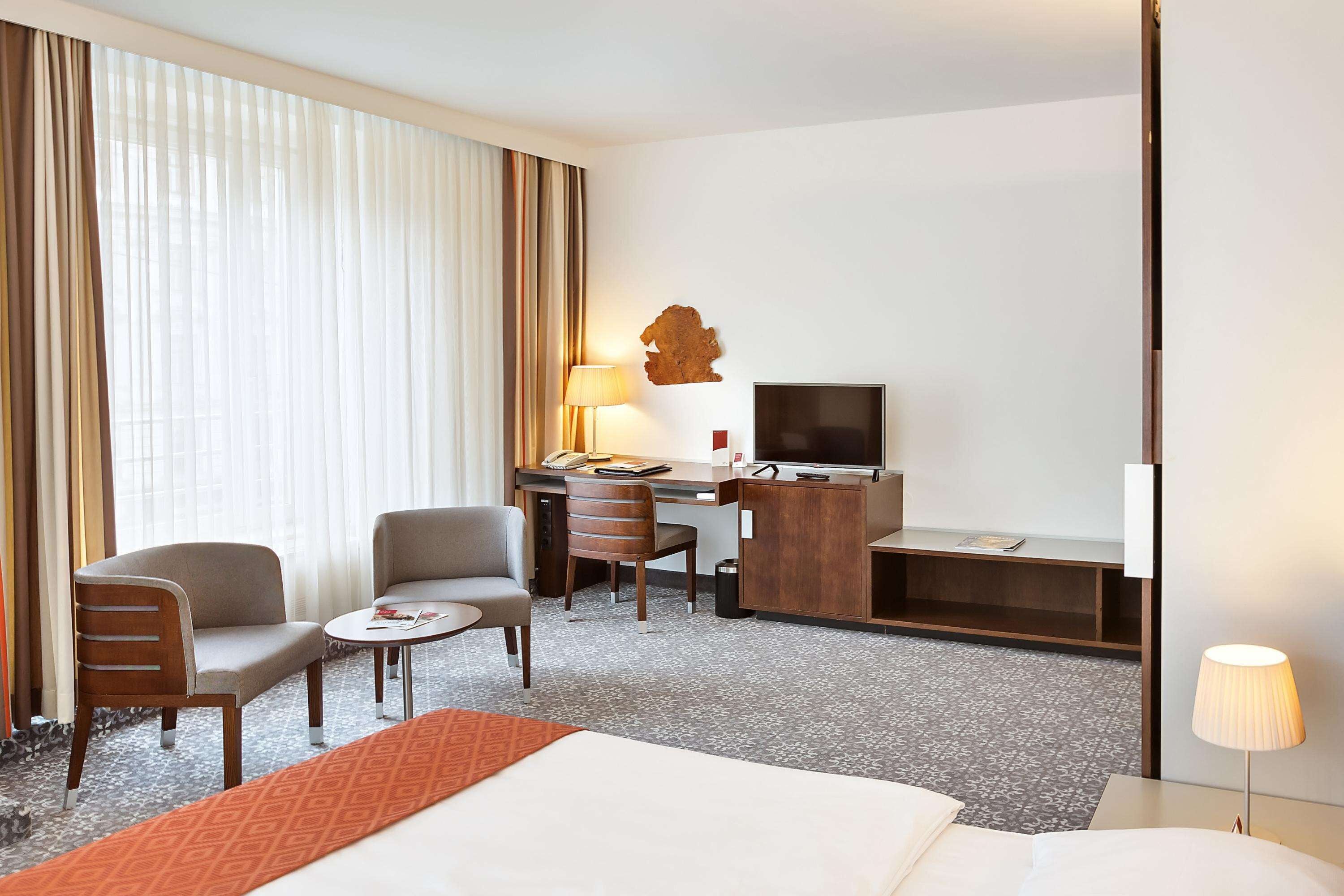 A hotel room with a bed, a desk, two chairs, a TV, and a lamp.