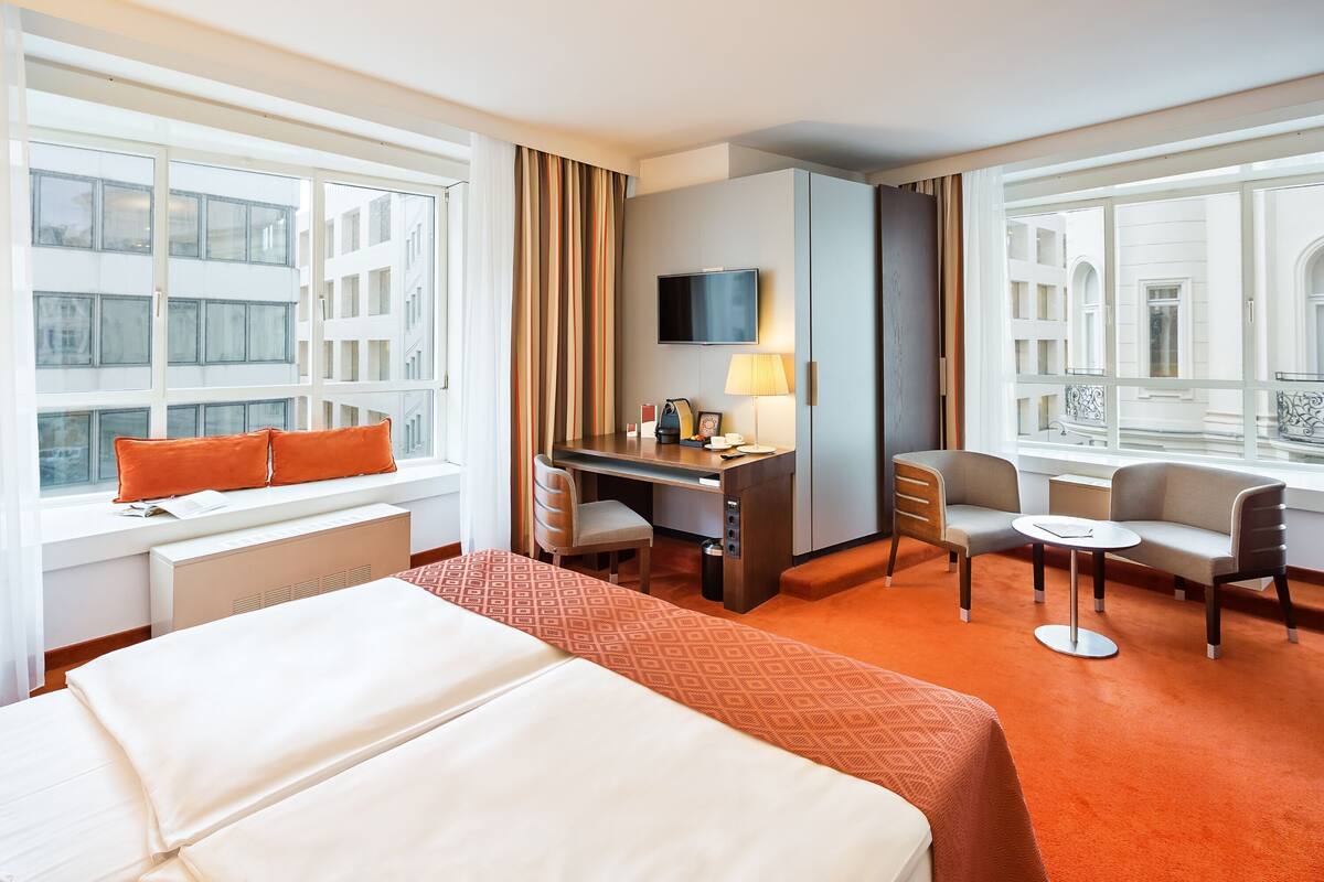 A hotel room with a bed, desk, chairs, a TV, and large windows.