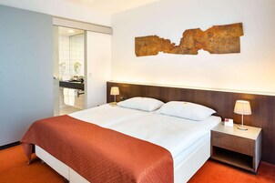 Quarto de hotel com uma cama grande, uma mesa de cabeceira com abajur, uma obra de arte na parede e um banheiro visível por uma porta aberta.