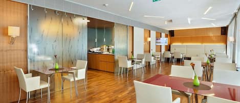 A modern hotel lobby with wooden flooring, white chairs, and tables. There is a reception desk with a TV and a kitchen area in the background.