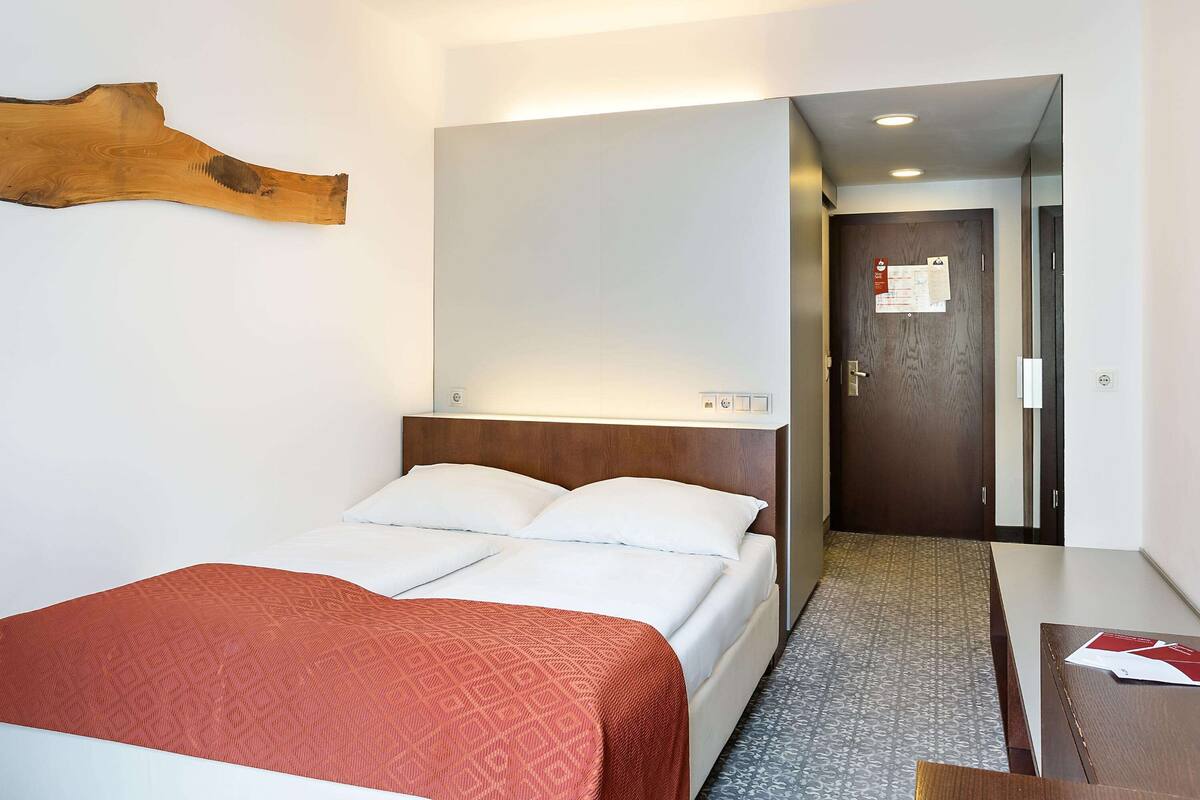 A hotel room with a bed, a wooden headboard, a red patterned bedspread, a wooden door, and a small table with papers.