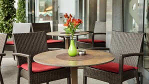 Una mesa redonda con sillas de mimbre y cojines rojos, un jarrón verde con flores y una pared de vidrio con señalización al fondo.