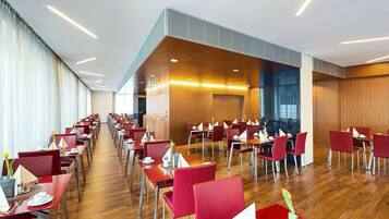A spacious dining area with red chairs and wooden tables, featuring a modern ceiling design and large windows.