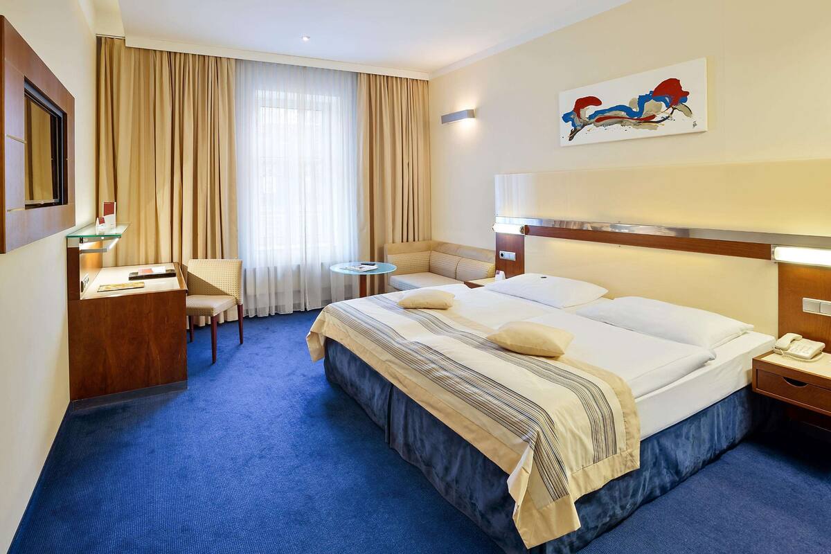 A hotel room with a large bed, a desk, a chair, a small table, and a painting on the wall.