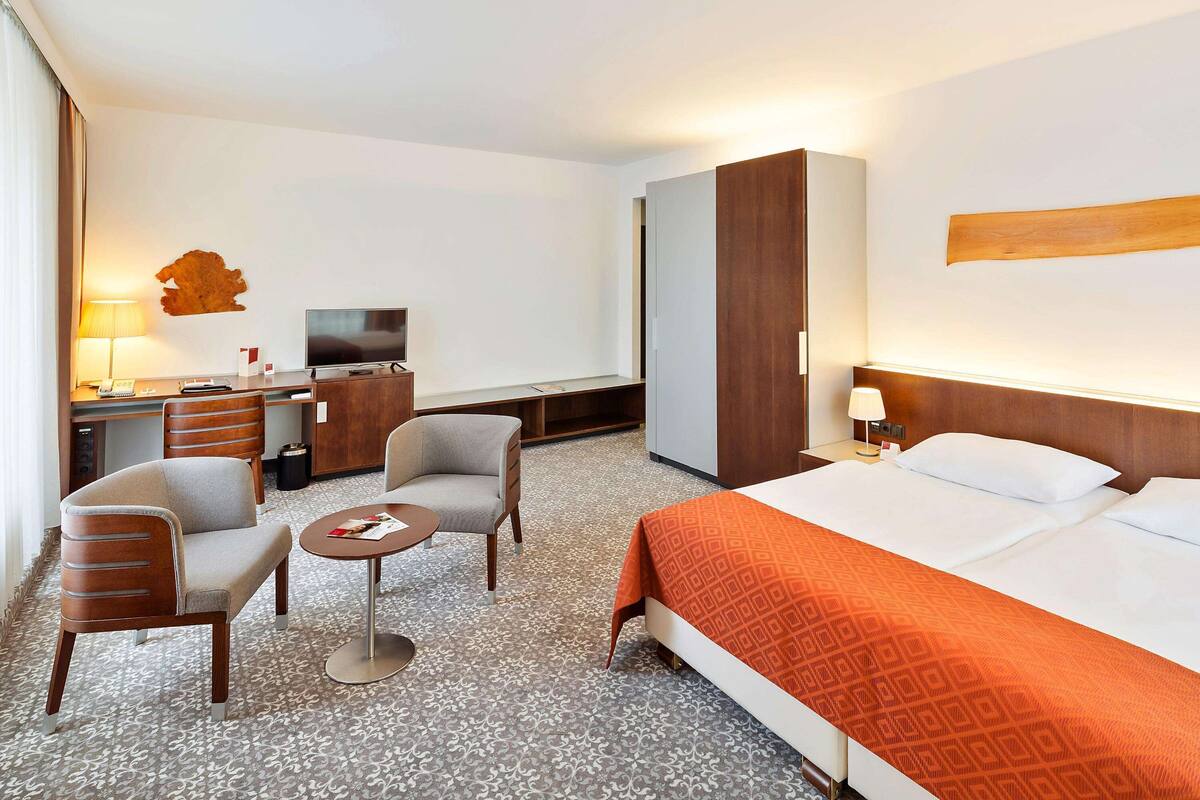 A hotel room with a bed, two armchairs, a small table, a TV, and a desk with a lamp.