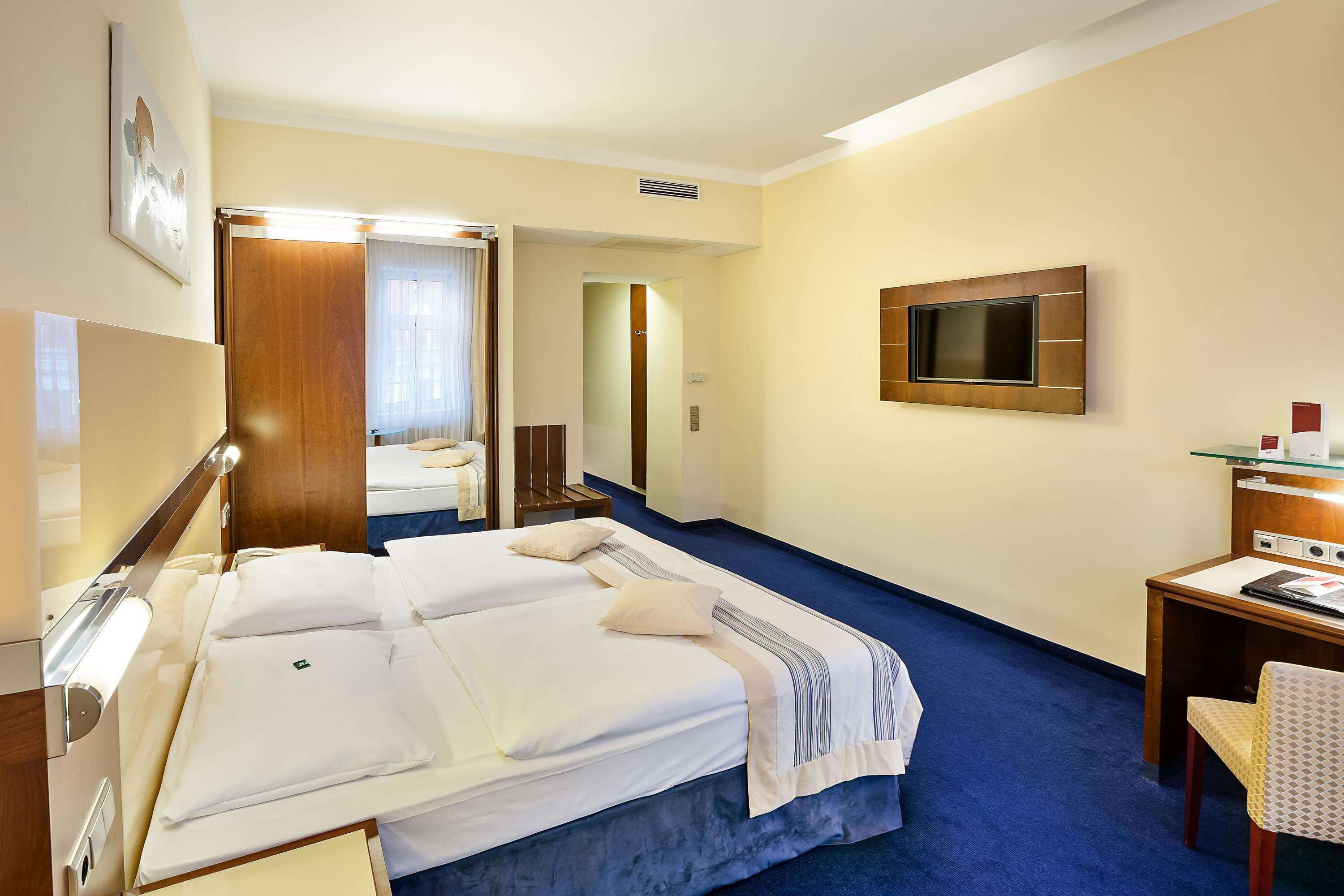 Quarto de hotel com uma cama grande, uma escrivaninha com cadeira, uma televisão na parede e um espelho.