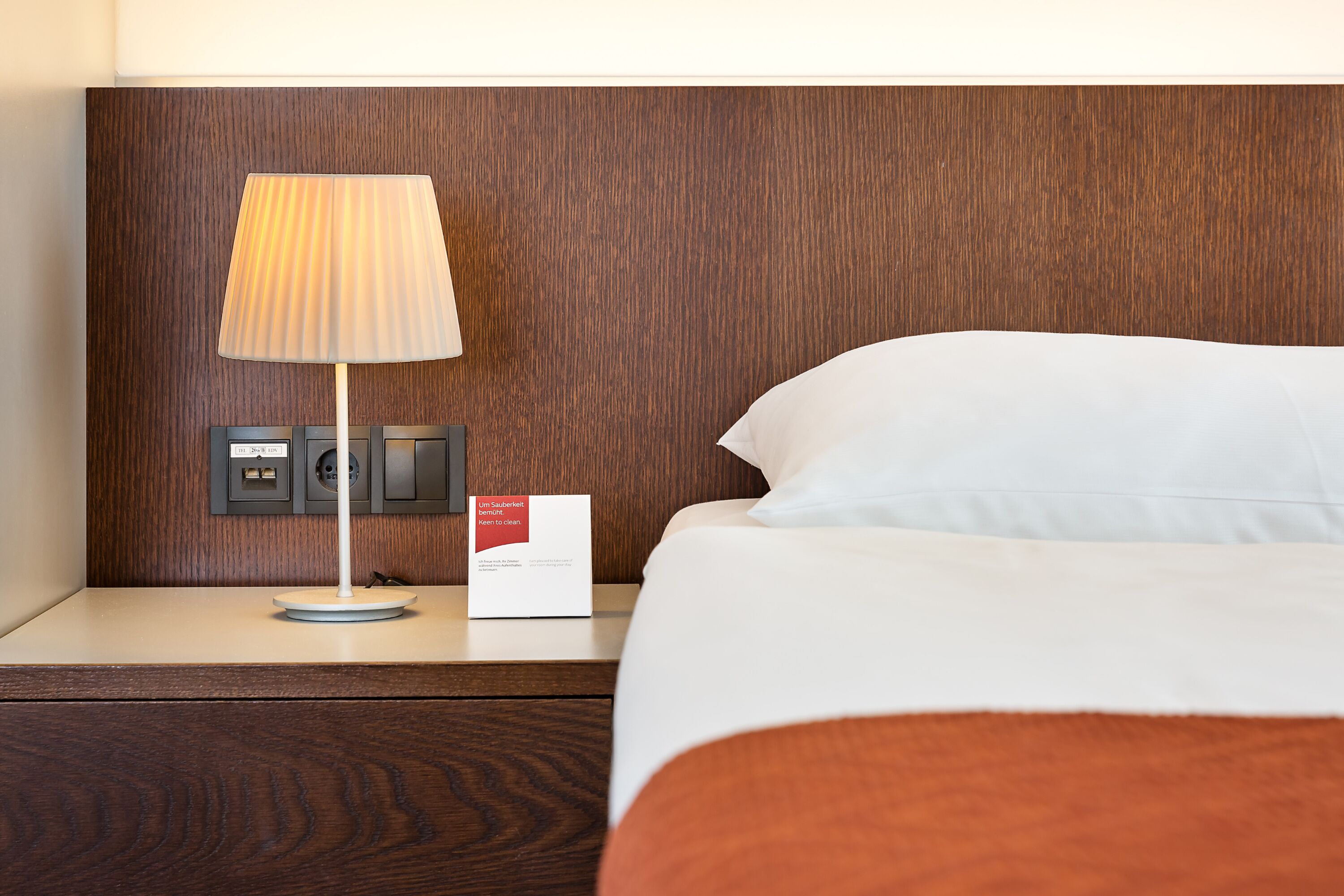 Cama bem arrumada com lençóis brancos, cabeceira de madeira, mesa de cabeceira com abajur e placa indicando os serviços do quarto.