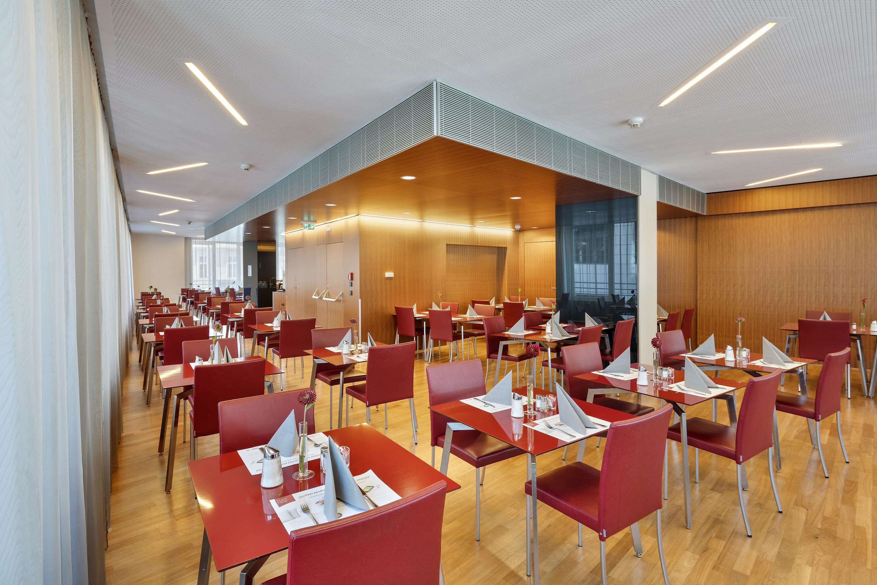A spacious dining area with red tables and chairs, a high ceiling with linear lighting, and a modern architectural design.
