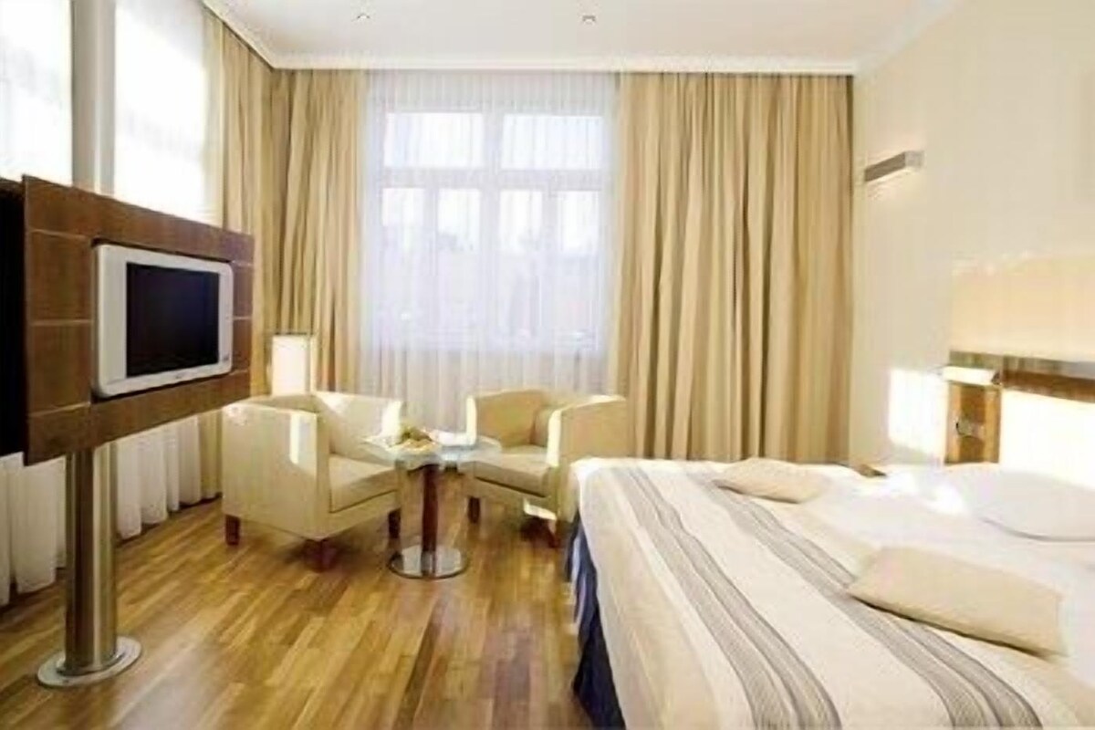A hotel room with a bed, two armchairs, a small table, a TV, and a window with curtains.