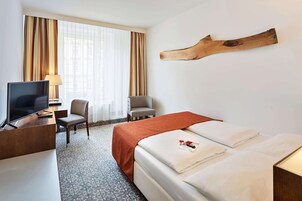 Quarto de hotel com cama, escrivaninha com televisão, cadeira e janela grande com cortinas.