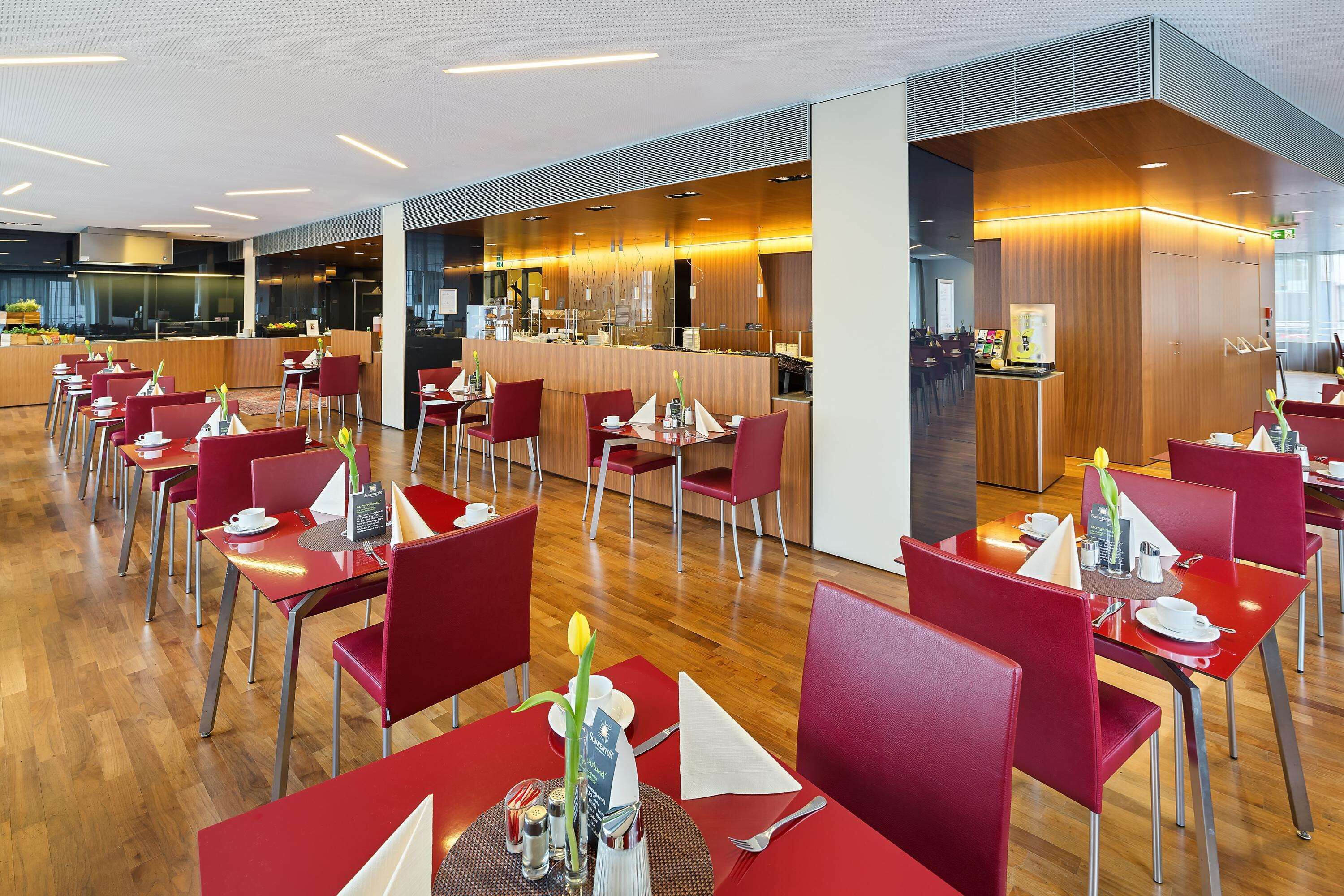 Um restaurante moderno com cadeiras e mesas vermelhas, piso de madeira e uma área de cozinha ao fundo.