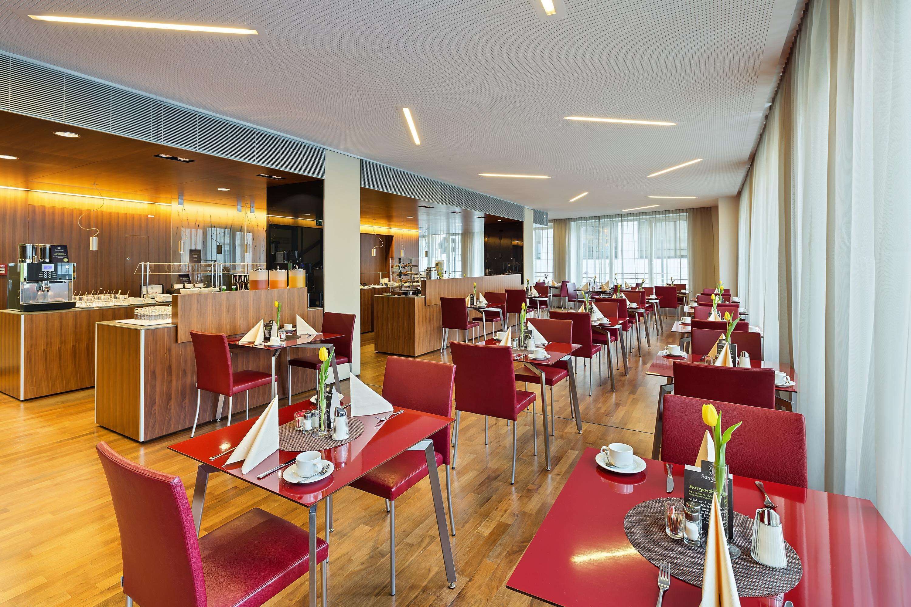 A modern dining area with red chairs and tables, a kitchen in the background, and large windows providing natural light.