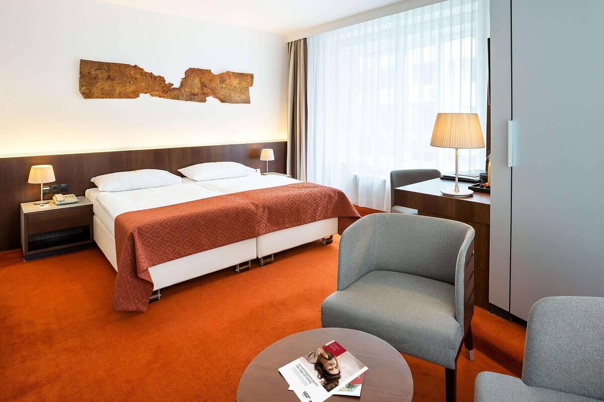 A hotel room with a large bed, two bedside tables with lamps, a desk with a chair, and a large window with curtains.