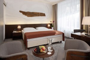 Quarto de hotel com uma cama grande, uma mesa de cabeceira com abajur, uma mesinha com frutas e um vaso, e uma cadeira.
