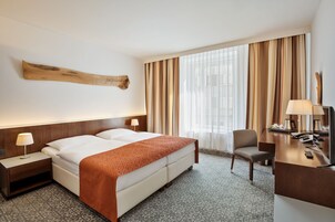 Quarto de hotel com uma cama grande, uma escrivaninha, uma cadeira, luminária de mesa, telefone e uma janela ampla com cortinas.