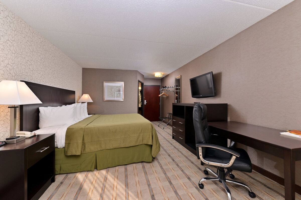 Quarto de hotel com cama, escrivaninha, cadeira, televisão e cabideiro.