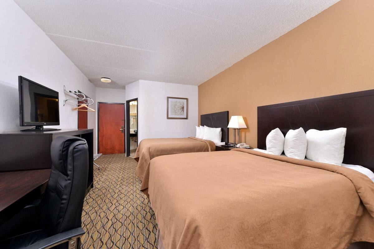 Quarto de hotel com duas camas, uma mesa de trabalho com televisão, uma cadeira e uma porta para um banheiro.