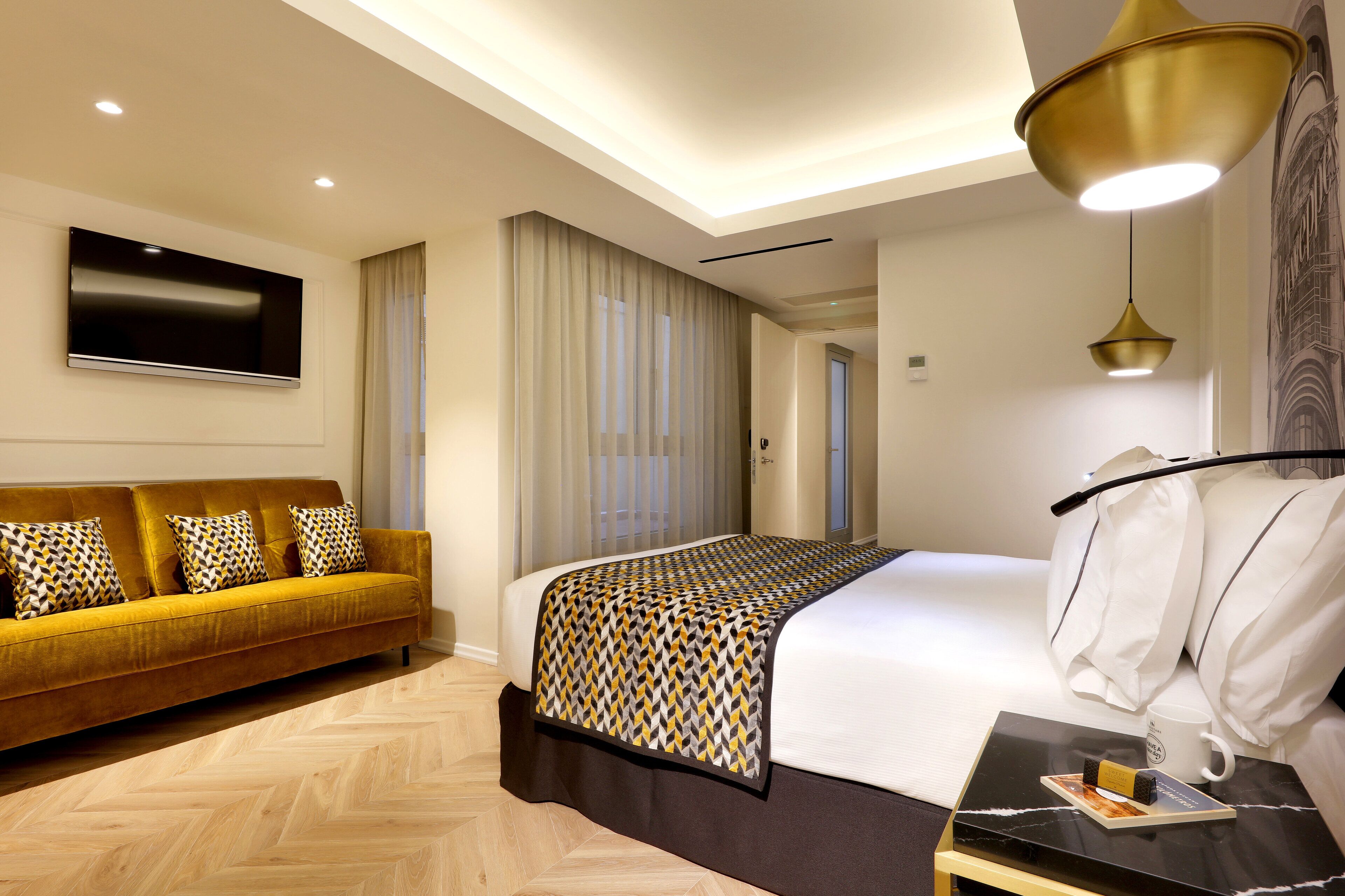 Habitación de hotel moderna con sofá, cama con ropa de cama estampada, mesita de noche con un libro y taza, y un ventanal con cortinas.
