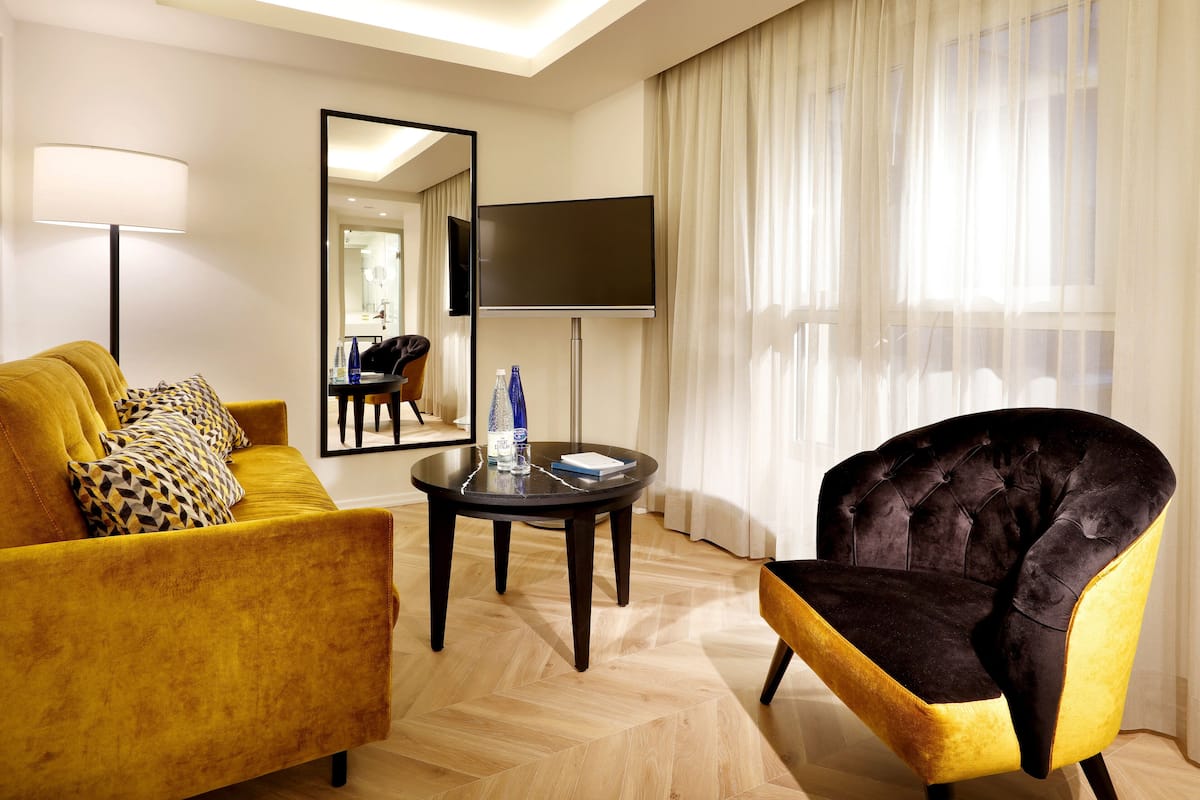 A hotel room with a yellow sofa, a black and gold armchair, a flat-screen TV, a small round table, and a large window with sheer curtains.