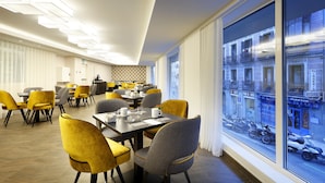 Un espace de restauration moderne avec des chaises et des tables jaunes, de grandes fenêtres offrant une vue sur la ville et un plafond au design géométrique.