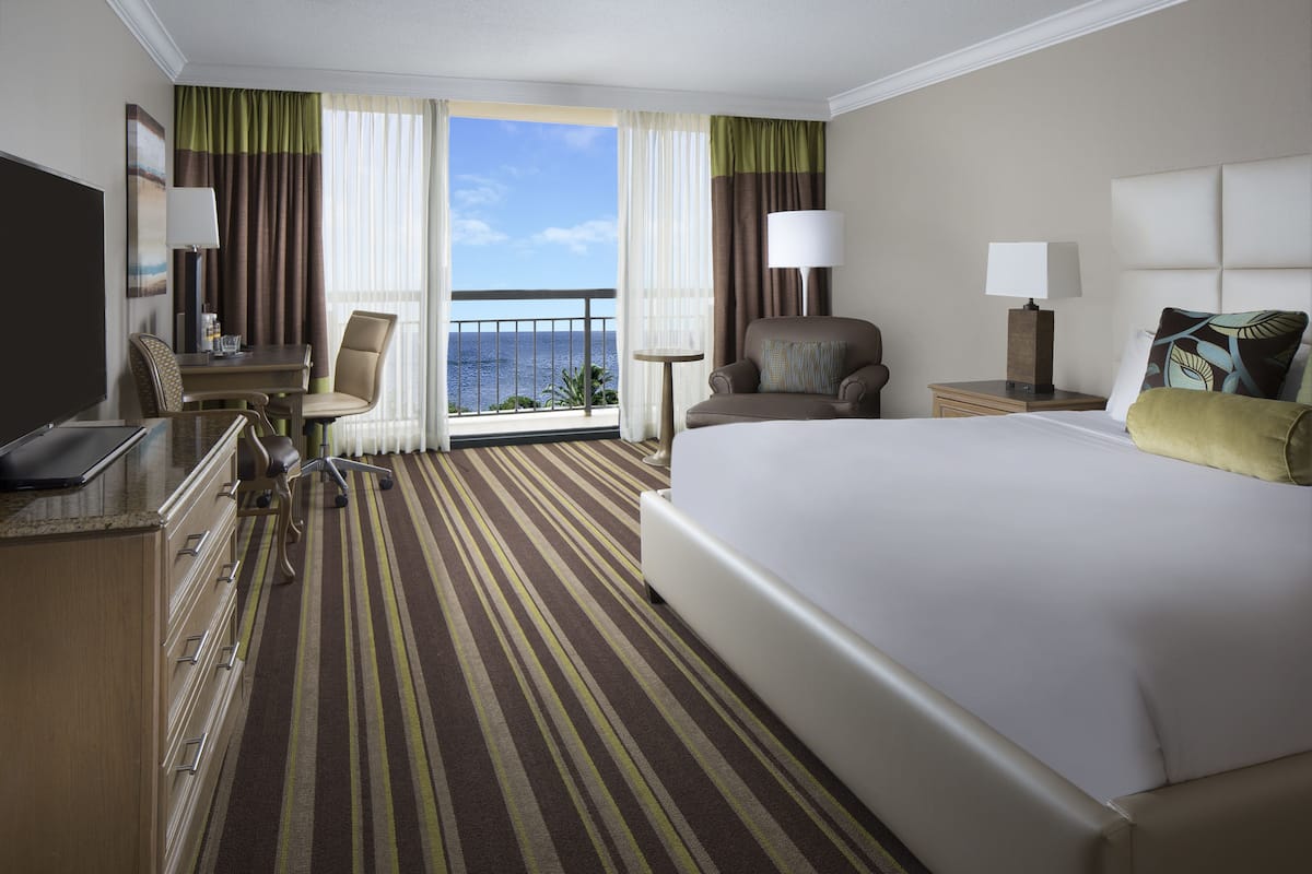A hotel room with a large bed, a desk with a chair, a small table, and a view of the ocean.
