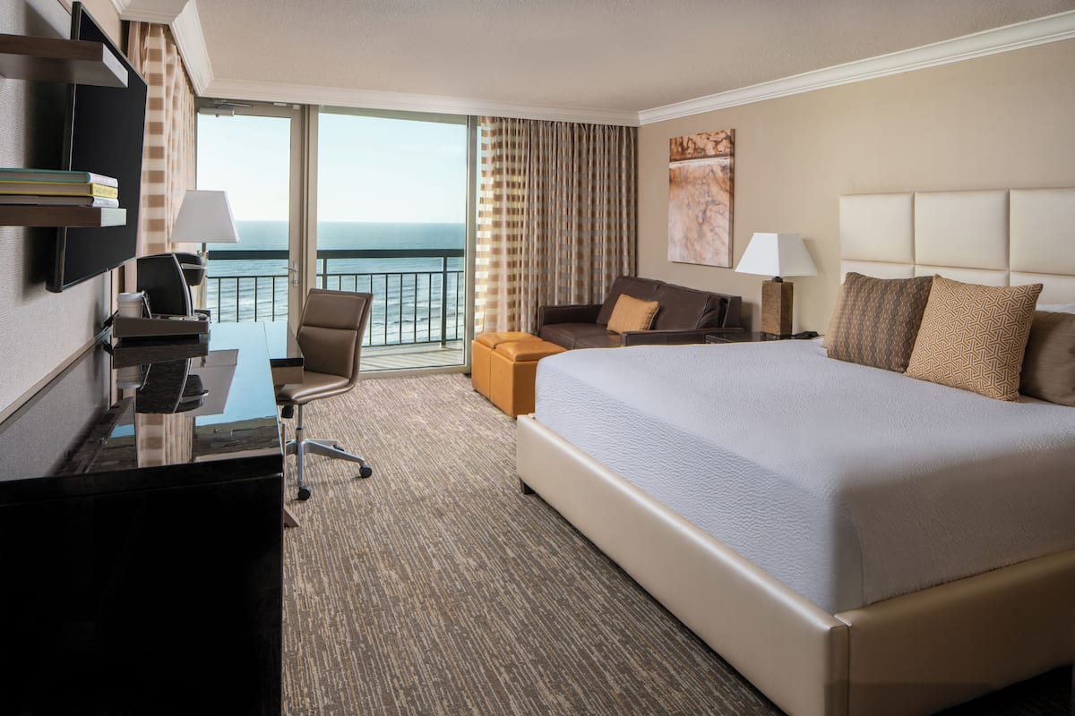 A hotel room with a large bed, a desk with a chair, a sofa, and a view of the ocean through large windows.