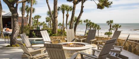 A poolside area with Adirondack chairs, a fire pit, and a view of the beach.