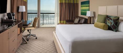 A hotel room with a large bed, a desk with a chair, a television, and a view of the ocean.
