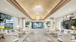 Eine geräumige Lobby mit einem prächtigen Kronleuchter, stilvoller Einrichtung und großen Fenstern mit Blick auf Palmen und ein Gebäude.