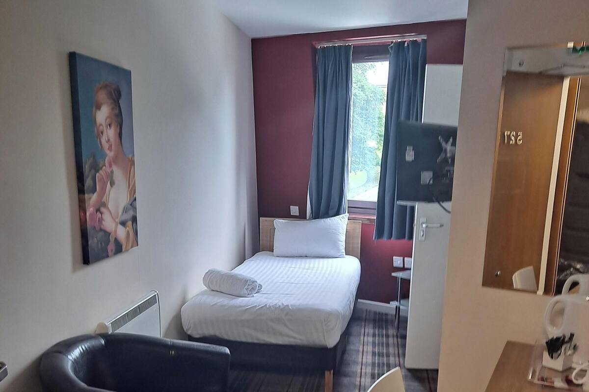 A hotel room with a bed, a black bathtub, a desk with a chair, a painting on the wall, and a window with curtains.