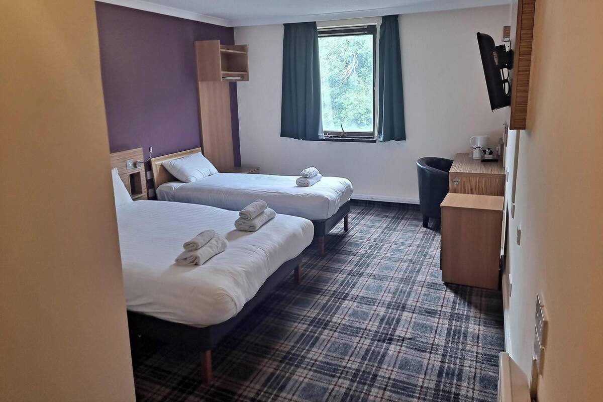 A hotel room with two beds, a small desk, a window with curtains, and a plaid-patterned carpet.