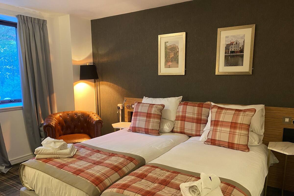 A hotel room with a double bed, two pillows, a chair, a lamp, and two framed pictures on the wall.
