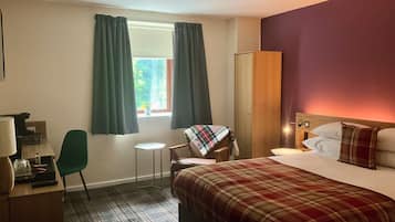 A hotel room with a bed, a desk, a chair, a small table, and a window with curtains.