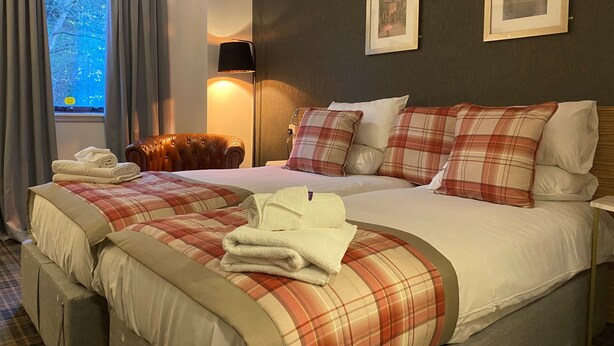 A hotel room with two beds, a large window with curtains, a leather armchair, and two framed pictures on the wall.