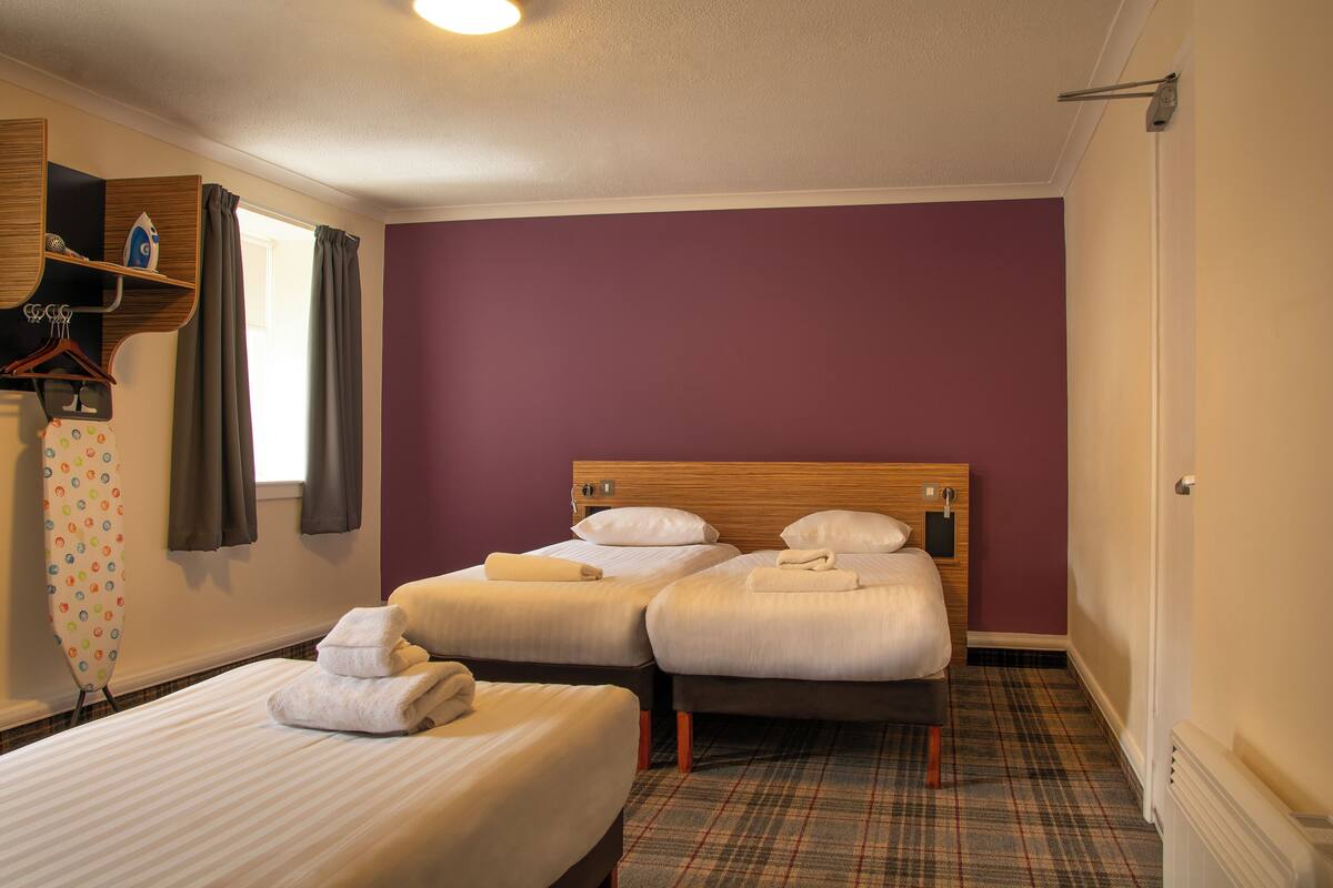 A hotel room with two beds, a wooden headboard, a plaid carpet, and a window with curtains.