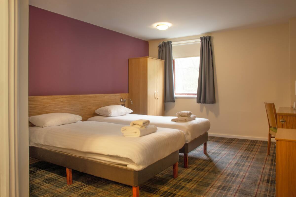 A hotel room with two beds, a wardrobe, a desk, and a window with curtains.