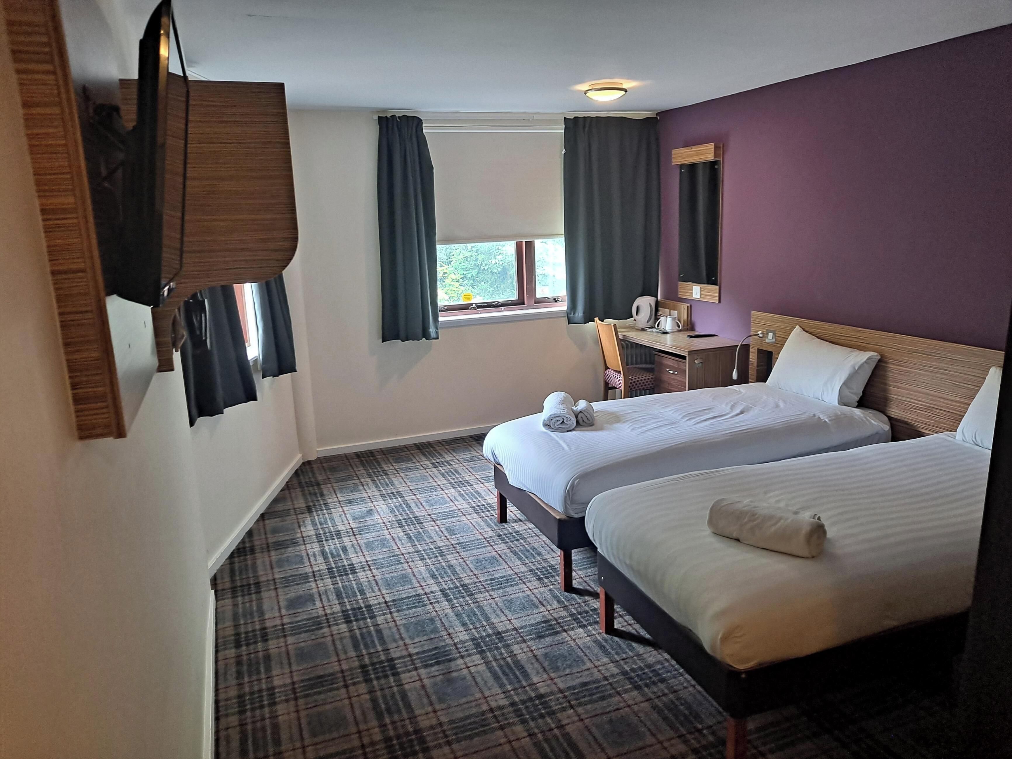 A hotel room with two beds, a desk, a chair, a window with curtains, and a mirror.