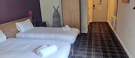 Habitación de hotel con dos camas, un armario de madera, alfombra de cuadros y un pasillo que conduce a otra habitación.