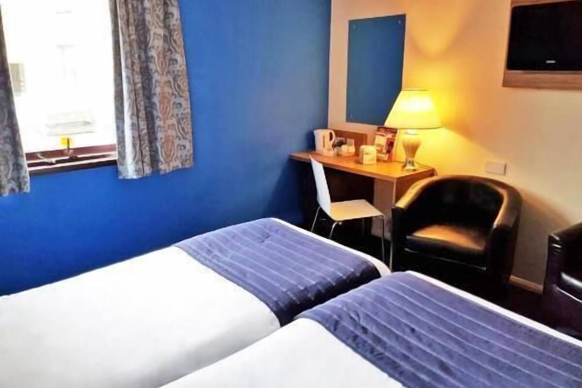 A hotel room with two beds, a desk, a chair, a TV, and a window with curtains.
