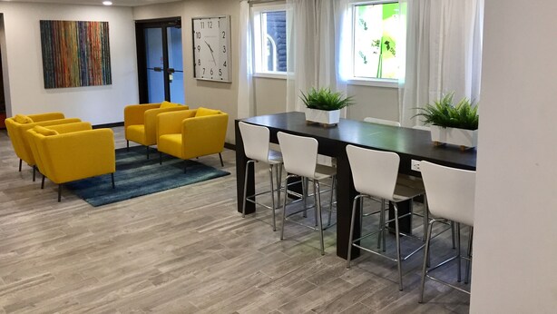 A modern dining area with a wooden table, white chairs, and yellow armchairs. There is a clock on the wall and a window with curtains.