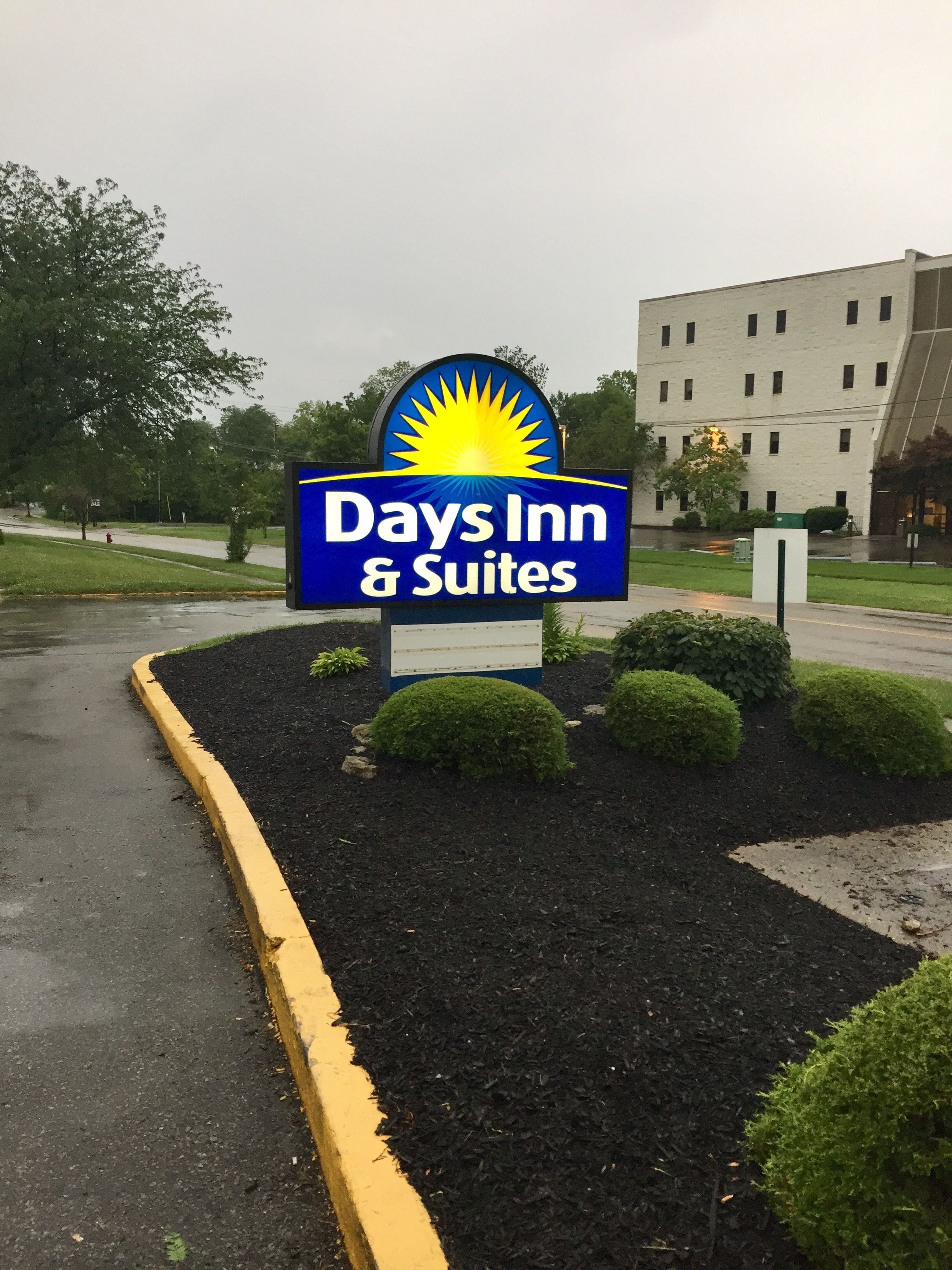 Señalización de Days Inn & Suites con un emblema de sol, rodeada de arbustos cuidados y una zona pavimentada.