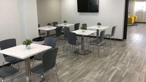 A modern conference room with white tables and grey chairs, equipped with potted plants and a flat-screen TV on the wall.