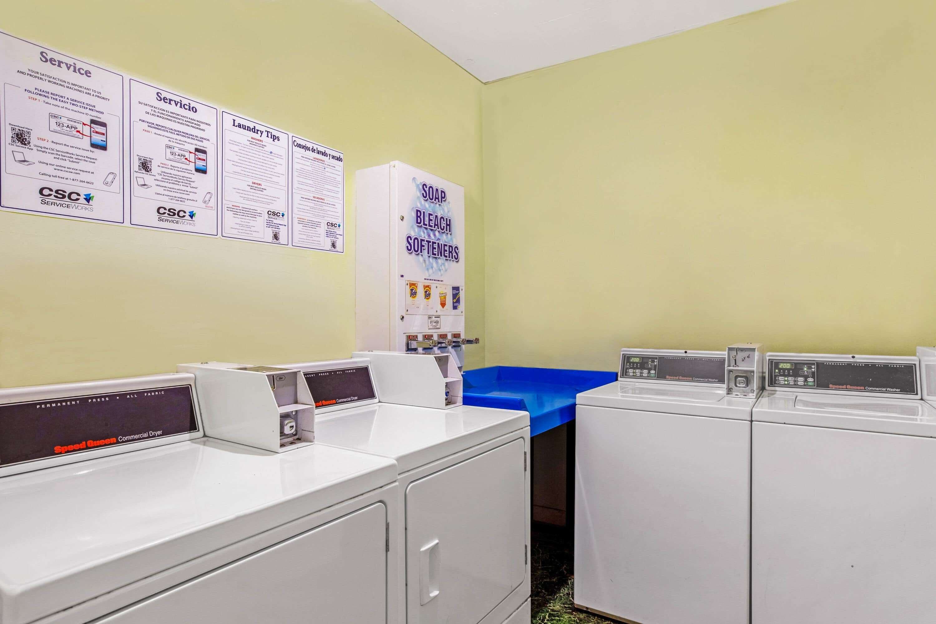 A laundry room with multiple washing machines, a blue drying rack, and informational posters on the wall.